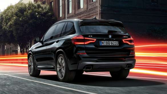 BMW X3 M40i sett skrått bakfra. Utvendig design på BMW X3 M40i i safirsort metallic som kjører, sett skrått bakfra i bylandskap med lysstriper.