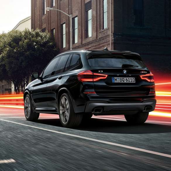 BMW X3 M40i sett skrått bakfra. Utvendig design på BMW X3 M40i i safirsort metallic som kjører, sett skrått bakfra i bylandskap med lysstriper.
