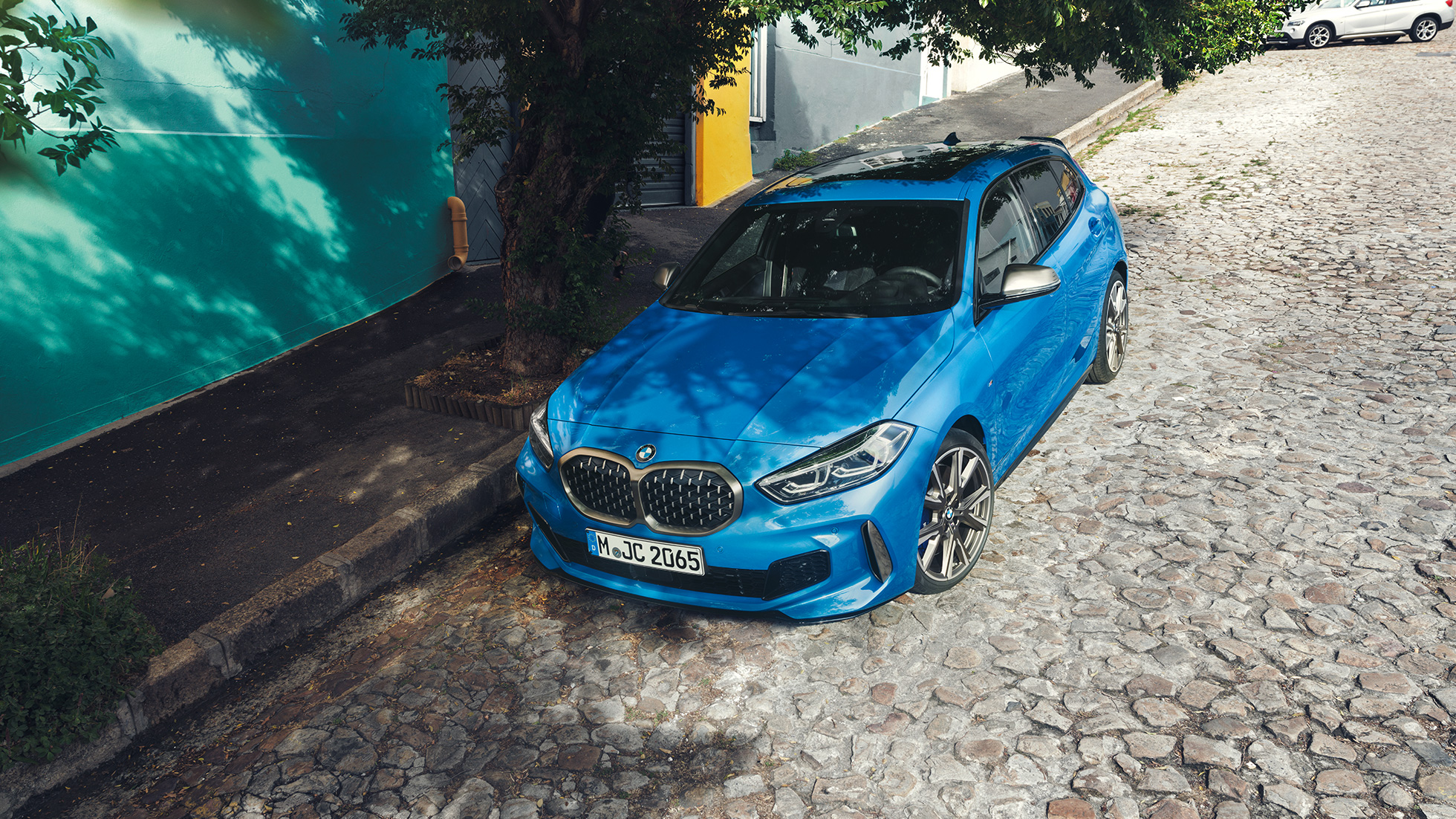 1-serie BMW på veikanten. 1-serie BMW i Misano Blau metallic, sett skrått forfra i fugleperspektiv, parkert i veikanten.