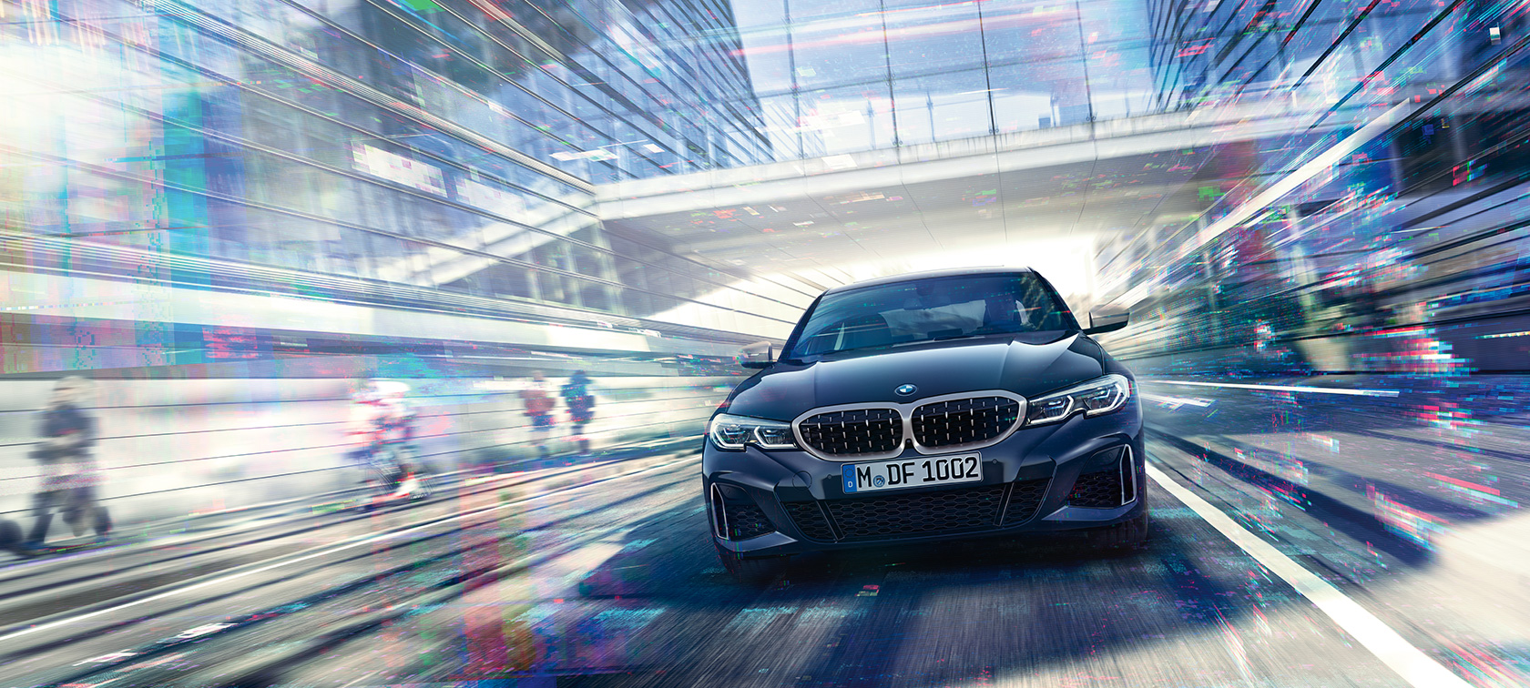 BMW 3-serie M340i xDrive Sedan sett forfra. BMW M340i xDrive Sedan som kjører mellom glassbygninger i byen.