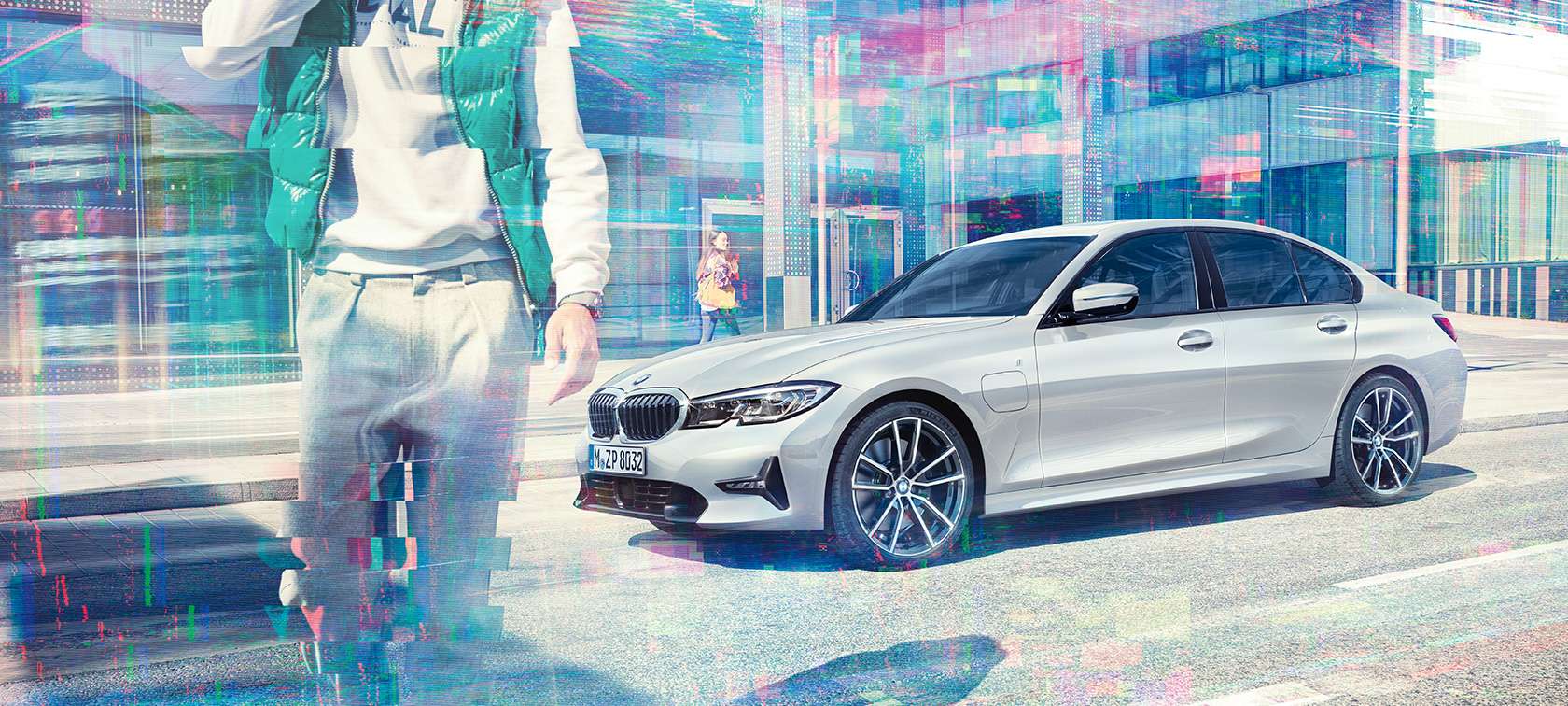 BMW 330e Sedan sett skrått fra siden foran glassbygning i byen med forbipasserende. BMW 3-serie 330e Sedan sett fra siden.
