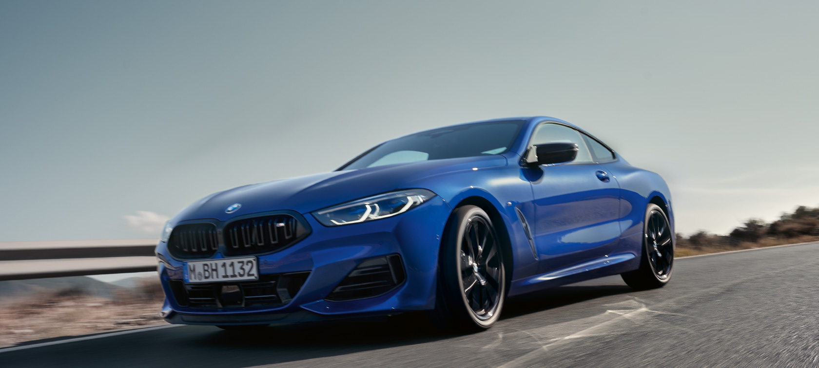 BMW 8-serie Coupé G15 LCI Facelift 2022 BMW M850i xDrive Portimaoblå sett forfra fra froskeperspektiv, kjørende BMW 8-serie Coupé G15 LCI Facelift 2022 BMW M850i xDrive Portimaoblå sett forfra fra froskeperspektiv, kjørende på en landevei foran et landskap med åser