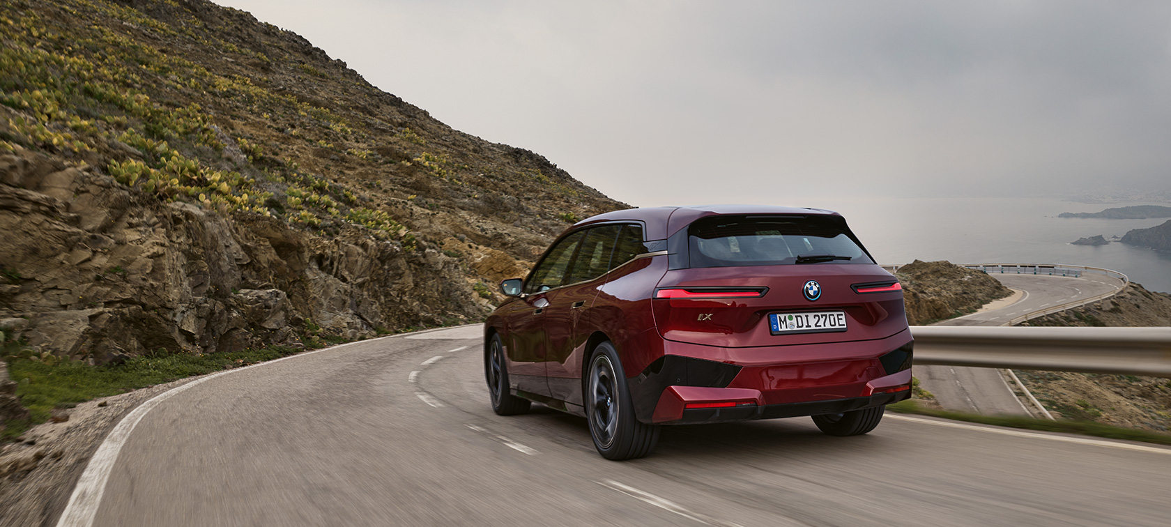 BMW iX i20 2021 elektrisk SUV BMW iX xDrive50 kjørende på kystvei BMW iX i20 2021 elektrisk SUV BMW iX xDrive50 Aventurinrød sett bakfra kjørende på kystvei