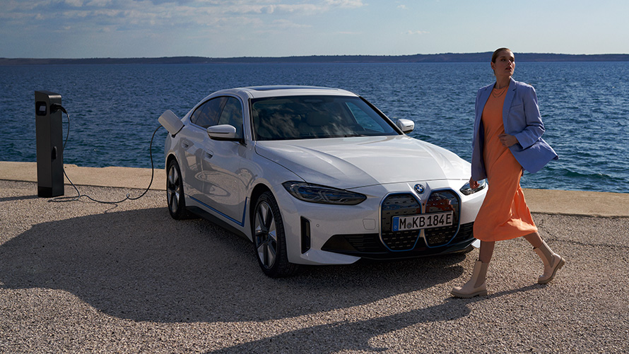 Sett skrått forfra BMW i4 eDrive40 G26 2021 Ved ladestasjon Kvinne foran BMW i4 eDrive40 G26 2021 Mineralhvit Sett skrått forfra ved ladesøyle foran innsjø