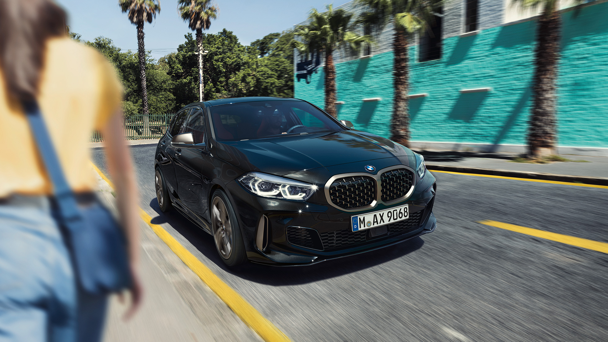 BMW M135i xDrive, sett skrått forfra. BMW M135i xDrive i safirsort metallic, sett skrått forfra, kjørende, med kvinne i forgrunnen.