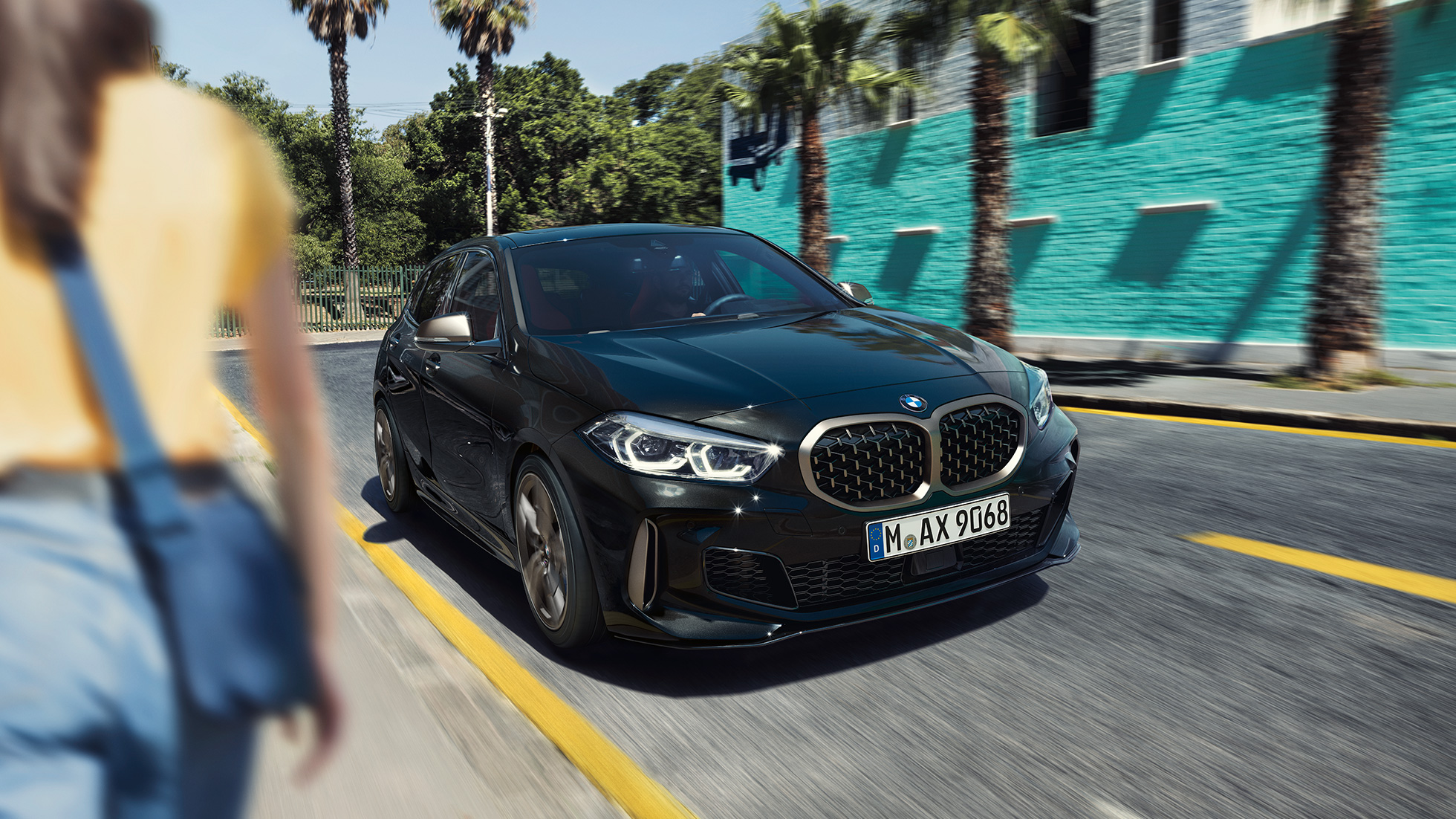 BMW M135i xDrive, sett skrått forfra. BMW M135i xDrive i safirsort metallic, sett skrått forfra, kjørende, med kvinne i forgrunnen.