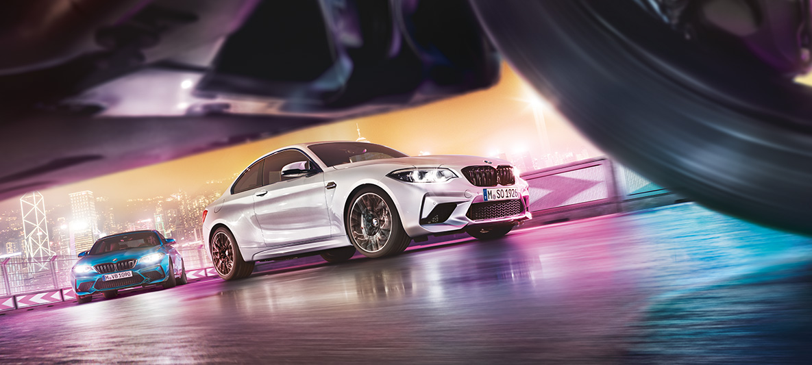BMW M2 Competition på reflekterende asfalt To BMW M2 Competition på banen sett på gatenivå foran urbant nattlandskap.