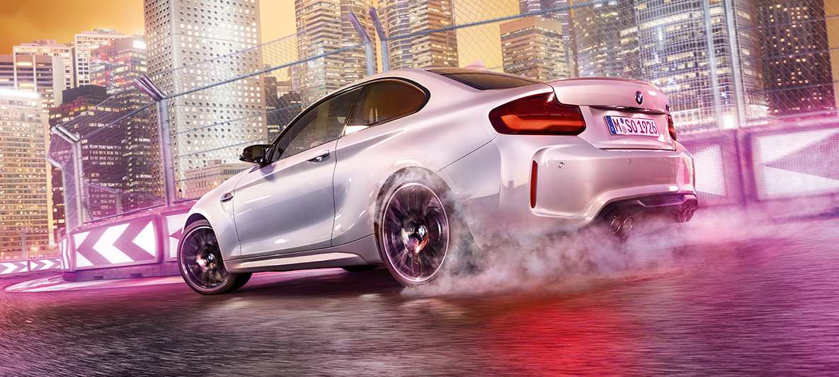 BMW M2 Competition gir gass Hvit BMW M2 Competition kjører på bane foran urban natthimmel.