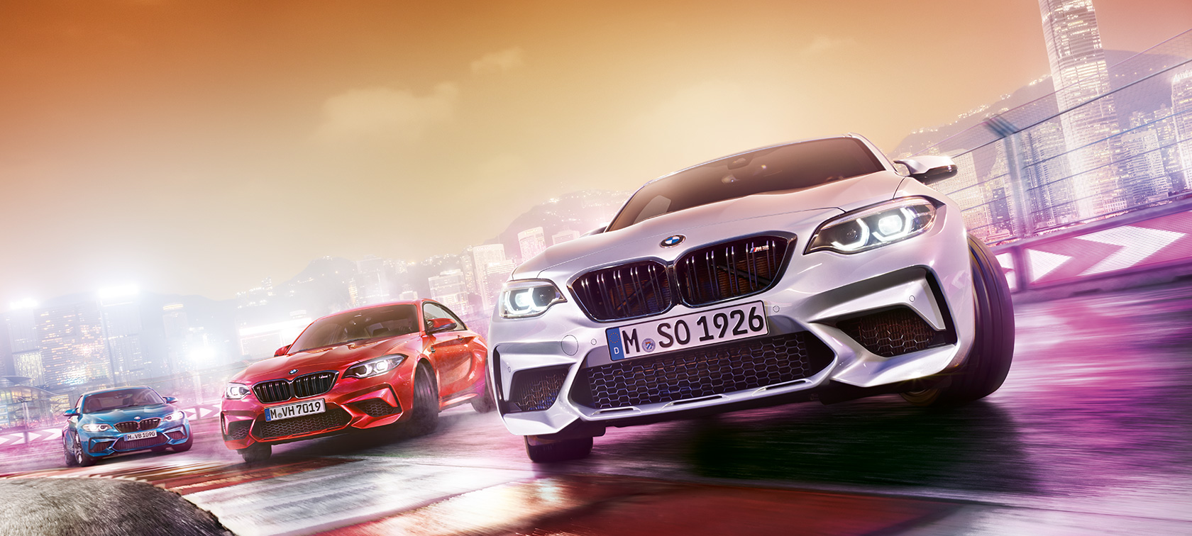 BMW M2 Competition i aksjon Tre BMW M2 Competition kjører på bane foran urbant nattlandskap.