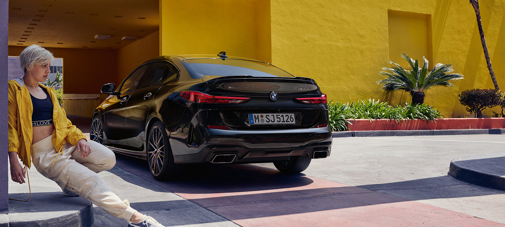 BMW M235i xDrive Gran Coupé sett skrått bakfra BMW M235i xDrive Gran Coupé Black safirsort metallic F44 sett skrått bakfra med dame i forgrunnen