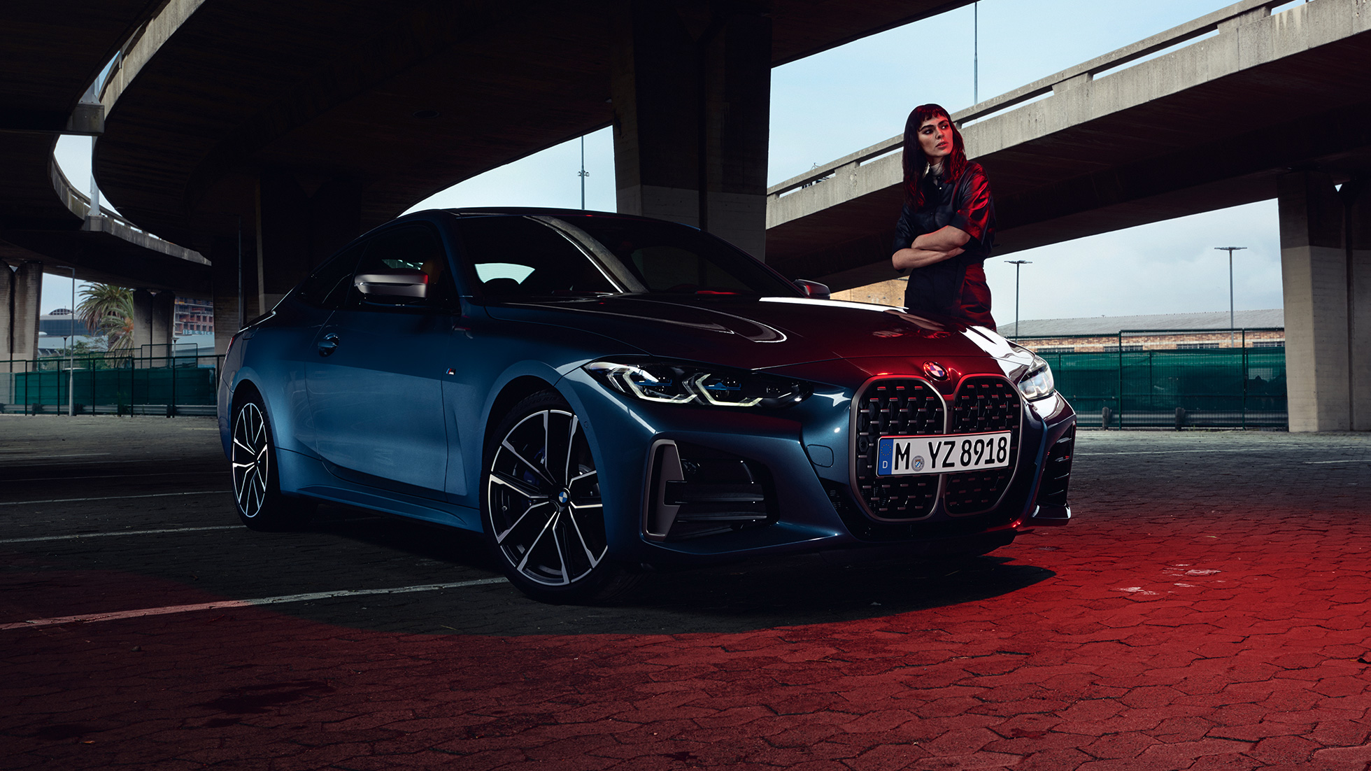 Modell står ved siden av BMW M440i xDrive Coupé G22 2020 Sett skrått fra siden BMW M440i xDrive Coupé Arctic Race blå metallic sett skrått fra siden Med modell