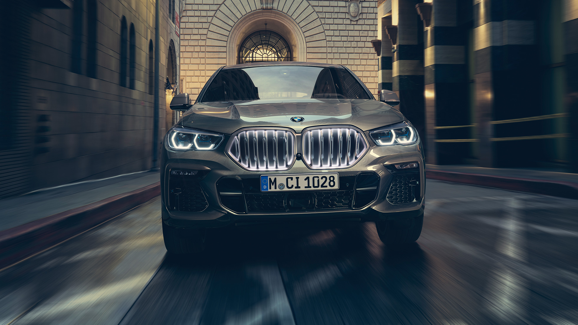 BMW X6 M50i med BMW ‚Iconic Glow‘-grill. BMW X6 M50i i frontperspektiv foran bylandskap.