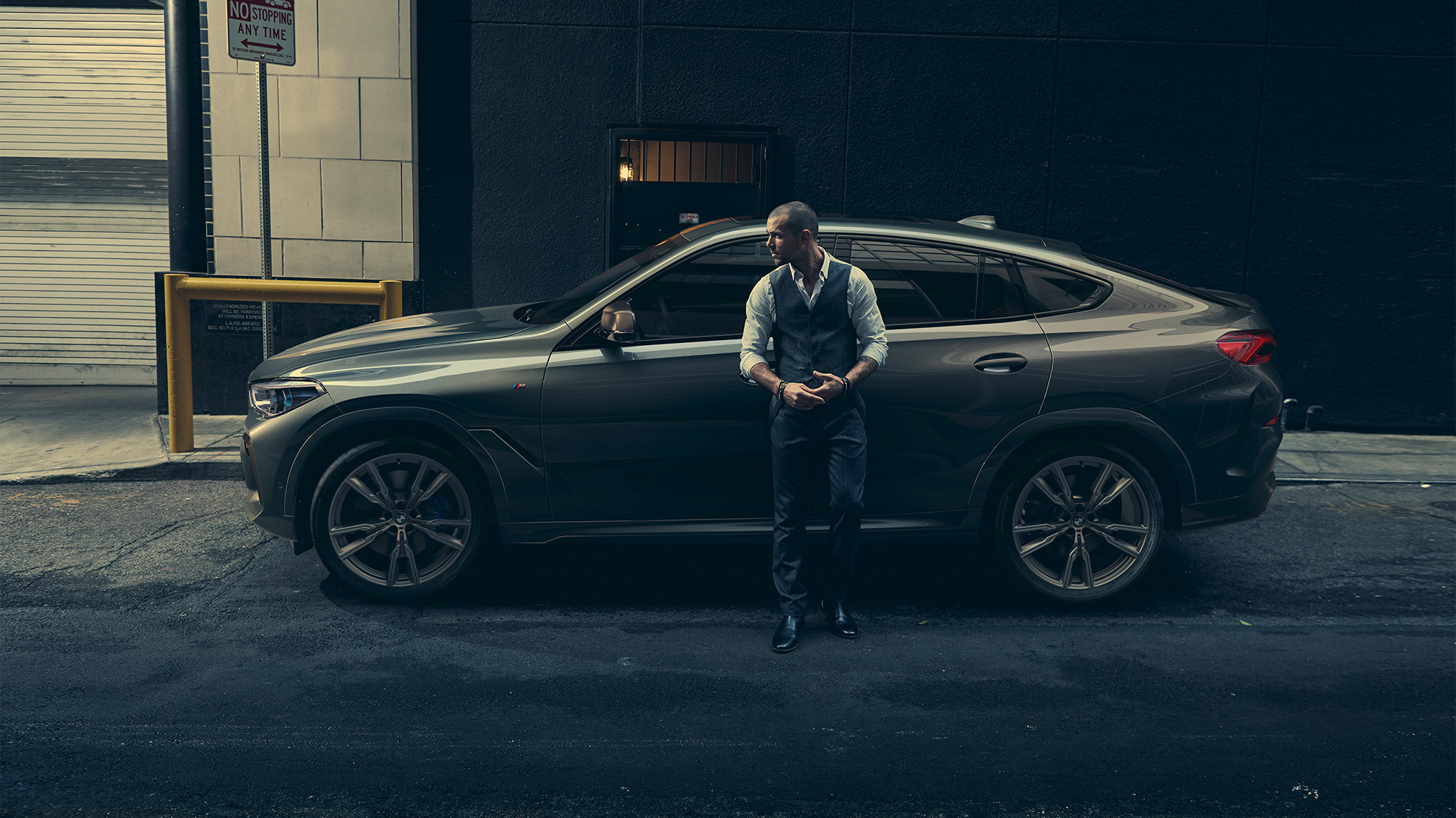 BMW X6 M50i sett fra siden. Mann lener seg mot BMW X6 M50i sett fra siden foran bylandskap.