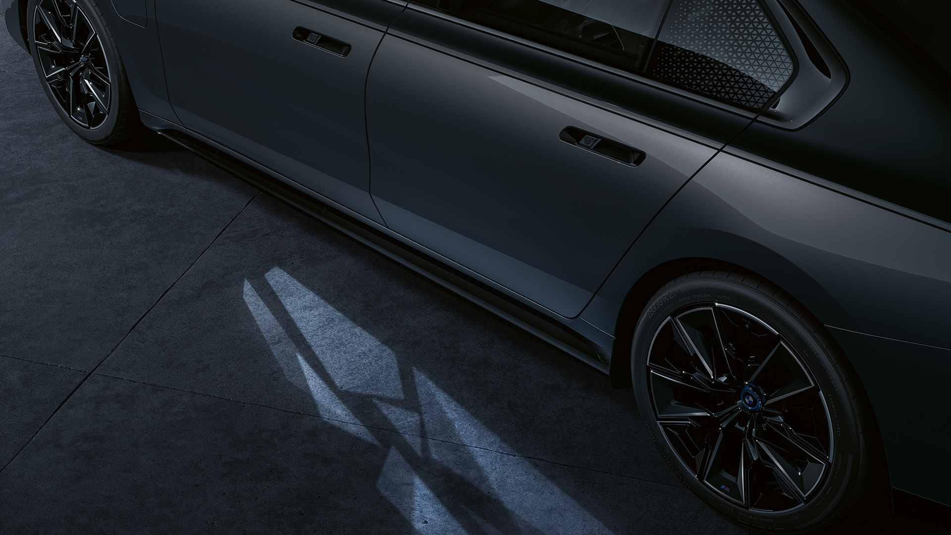 Lyskonsept på BMW M760e xDrive Sedan G70 Lyskonseptet BMW M760e xDrive Sedan G70 Two-Tone-lakkering safirsort metallic BMW Individual dravittgrå metallic sett fra siden med nærbilde av tent Welcome Light Carpet