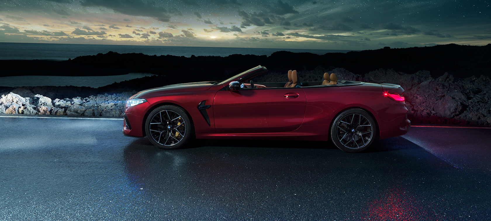 BMW M8 Competition Cabriolet, sett fra siden. BMW M8 Competition Cabriolet i Motegi rød metallic med kalesjen nede, sett fra siden, stående på en kystvei under stjernehimmel.