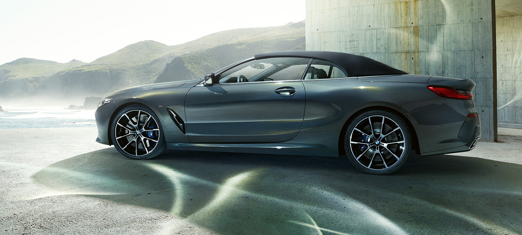 BMW M850i xDrive Cabriolet, sett fra siden. BMW M850i xDrive Cabriolet i dravittgrå metallic med kalesjen oppe, sett fra siden foran fjellkjede.