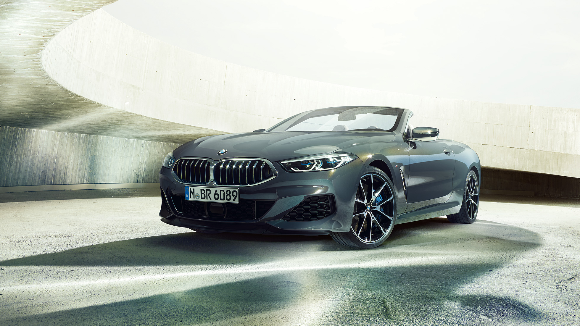 BMW M850i xDrive Cabriolet, sett skrått forfra. BMW M850i xDrive Cabriolet i dravittgrå metallic med kalesjen nede, sett skrått forfra, stående i betongbygning.