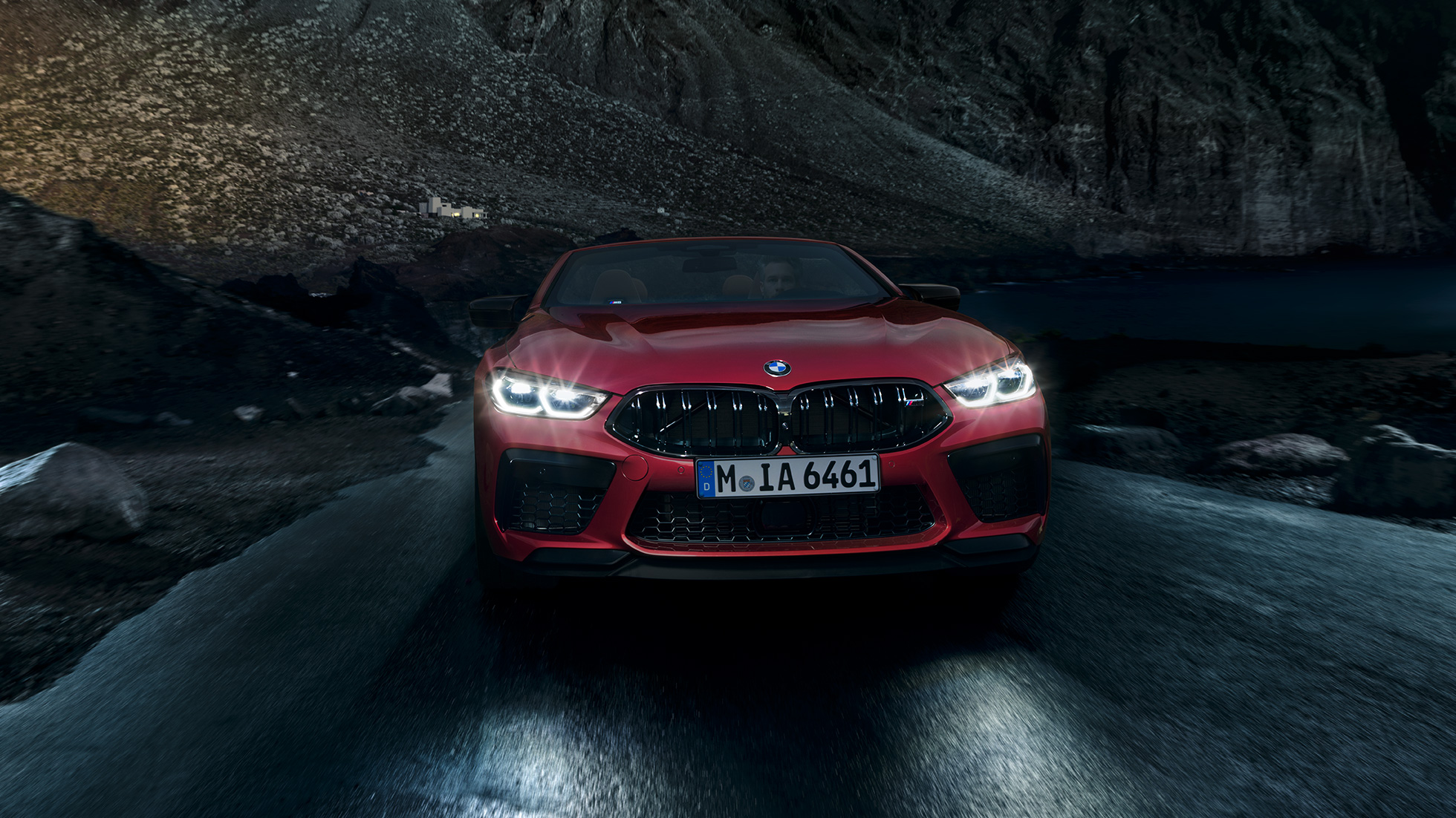 BMW M8 Competition Cabriolet, sett forfra. BMW M8 Competition Cabriolet i Motegi rød metallic med frontlyktene på, sett forfra kjørende om natten.