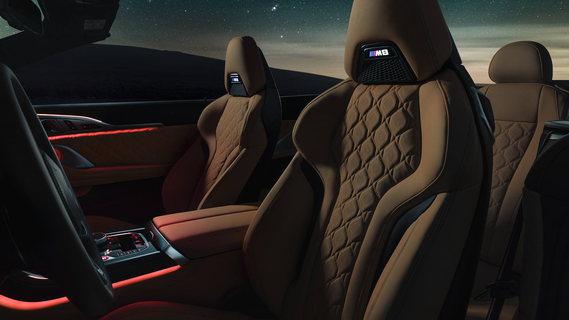 M sportsseter i merinoskinn. BMW M8 Competition Cabriolet med kalesjen oppe under stjernehimmel, interiør med M sportsseter i merinoskinn og rød bakgrunnsbelysning.