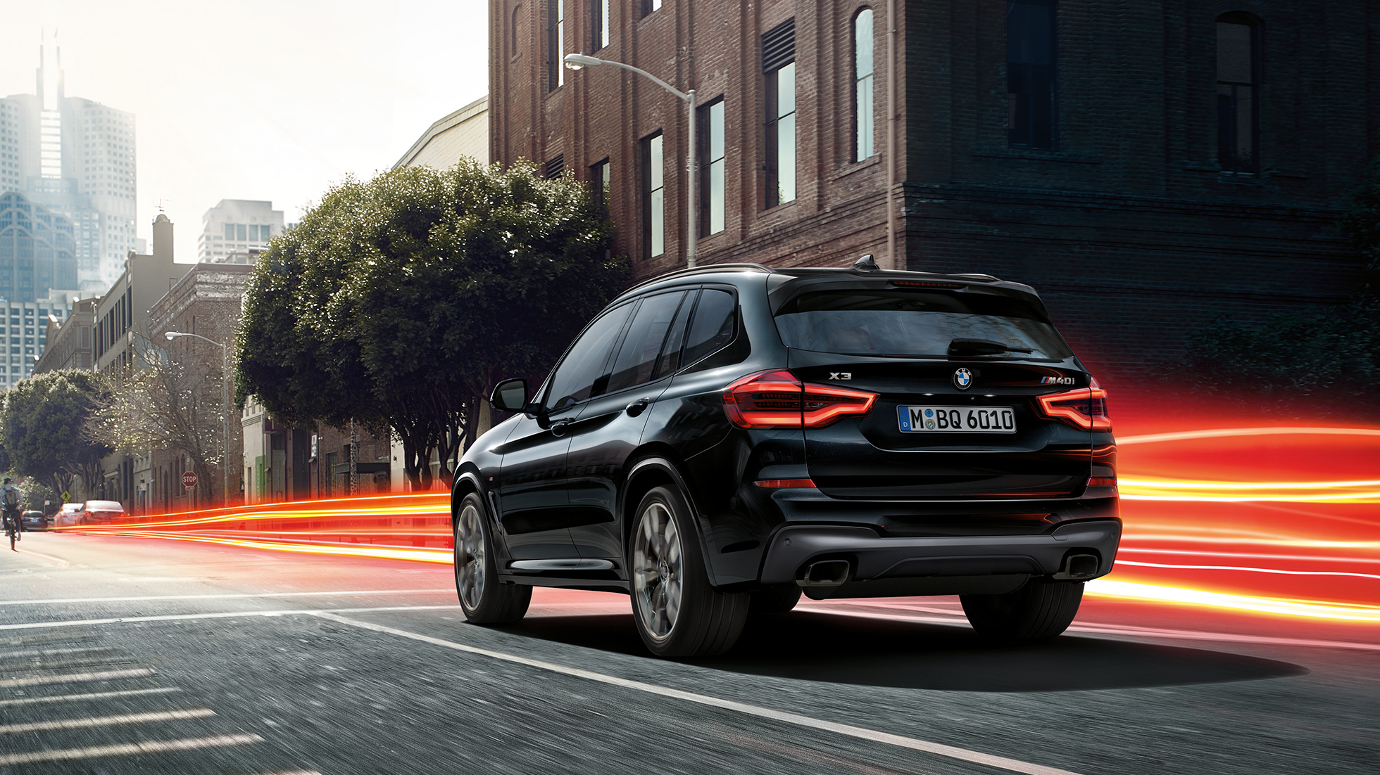BMW X3 M40i sett skrått bakfra. Utvendig design på BMW X3 M40i i safirsort metallic som kjører, sett skrått bakfra i bylandskap med lysstriper.