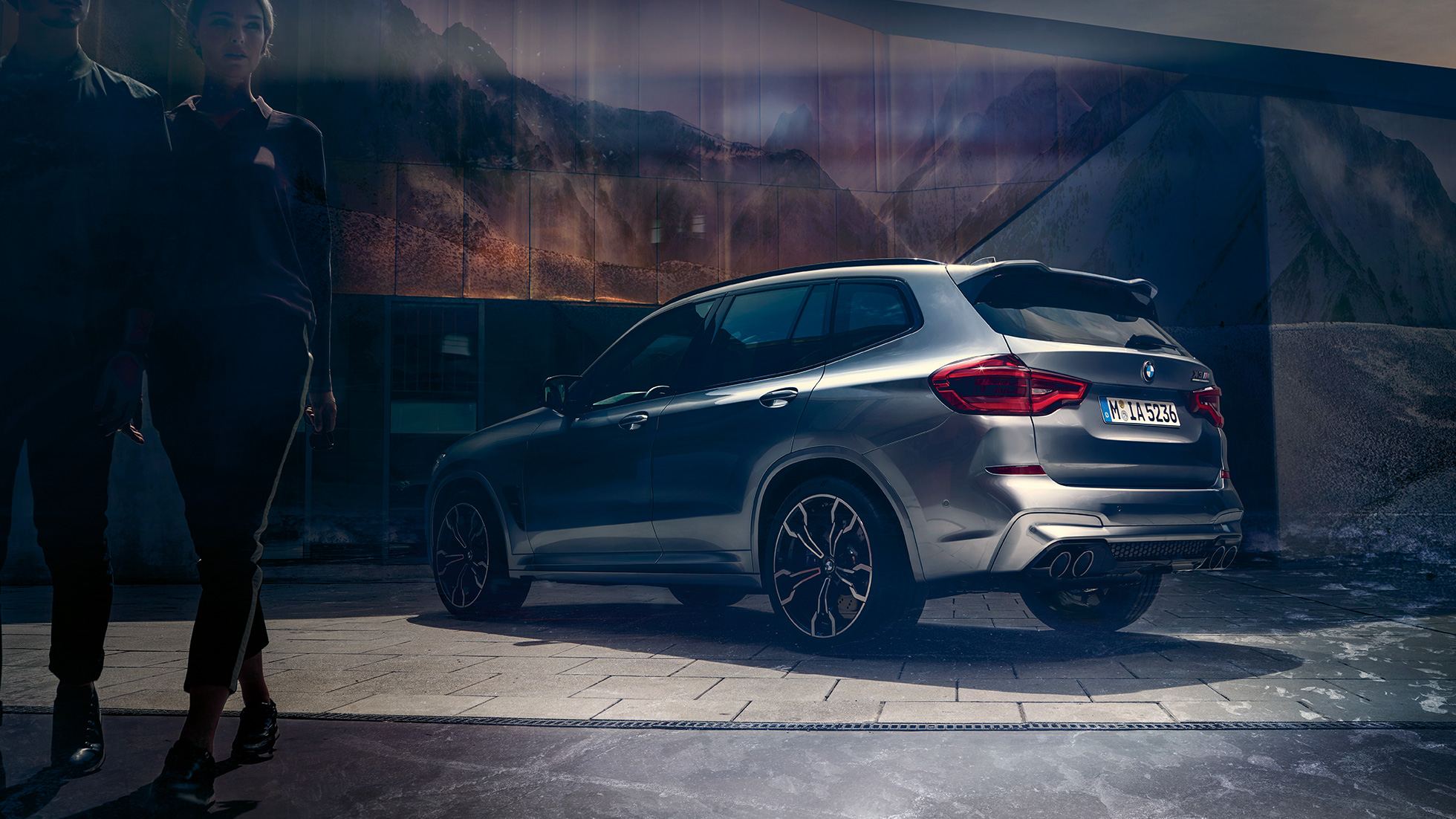 BMW X3 M Competition sett fra siden bak. Utvendig design på BMW X3 M Competition i doningtongrå metallic med to personer i forgrunnen sett fra siden bak.