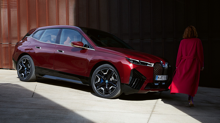 Kvinne foran BMW iX i20 2021 sett skrått forfra Kvinne i rød frakk foran BMW iX i20 2021 sett skrått forfra