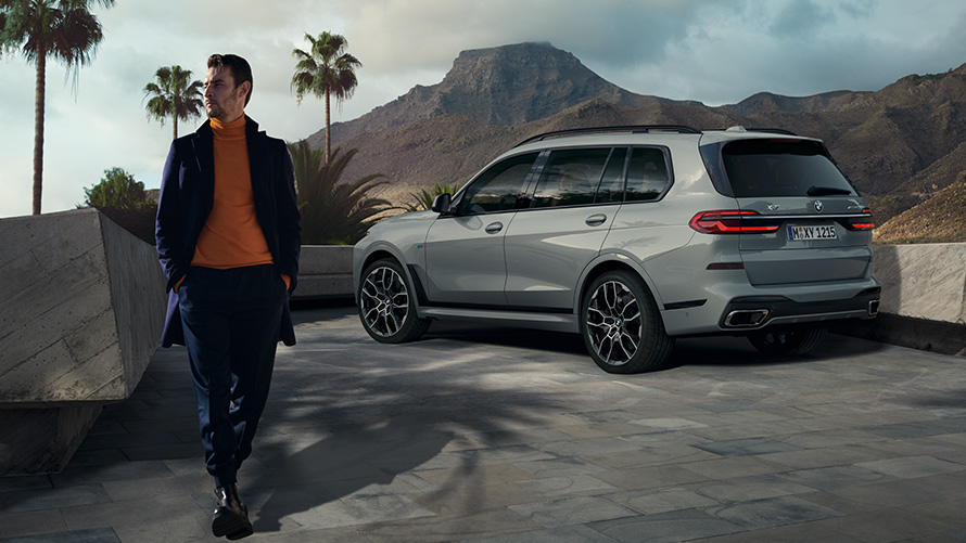Mann foran parkert BMW X7 (G07) Remote Control Parking Remote Control Parking Mann foran parkert BMW X7 (G07) Sett skrått bakfra under palmer med fjellandskap i bakgrunnen