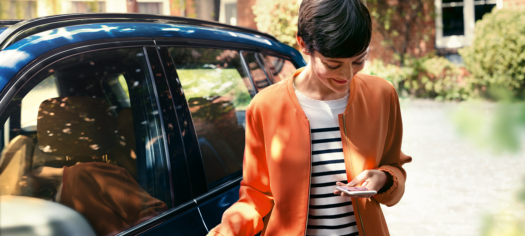 Mørkhåret kvinne foran en BMW Mørkhåret kvinne i oransje jakke foran en BMW ser på smarttelefonen