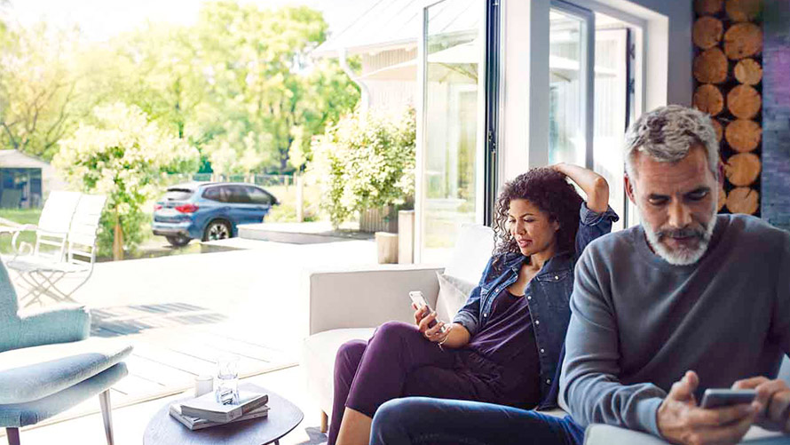 BMW Remote Services En mann og en kvinne sitter i sofaen og bruker Remote Services på smarttelefonene sine