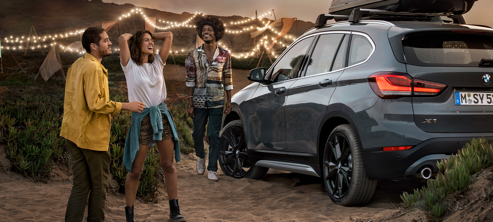 jublende trio i BMW X1 med BMW tilbud sommer Lykkelig trio i mineralgrå BMW X1 utstyrt med BMW tilbud sommer, sett skrått bakfra ved stranden