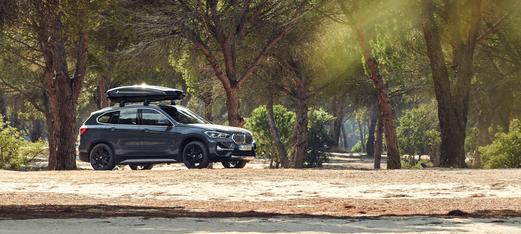 Sett fra siden BMW X1 med takboks BMW tilbud sommer Sett fra siden BMW X1 mineralgrå med BMW takboks stående offroad i skogen BMW tilbud sommer