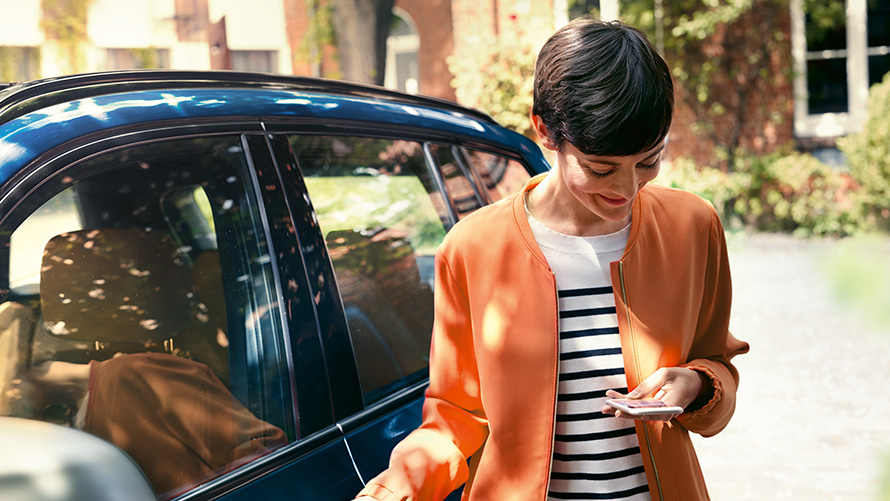BMW tilbud sommer BMW Teleservices BMW tilbud sommer BMW Teleservices dame står foran BMW og bruker en smarttelefon