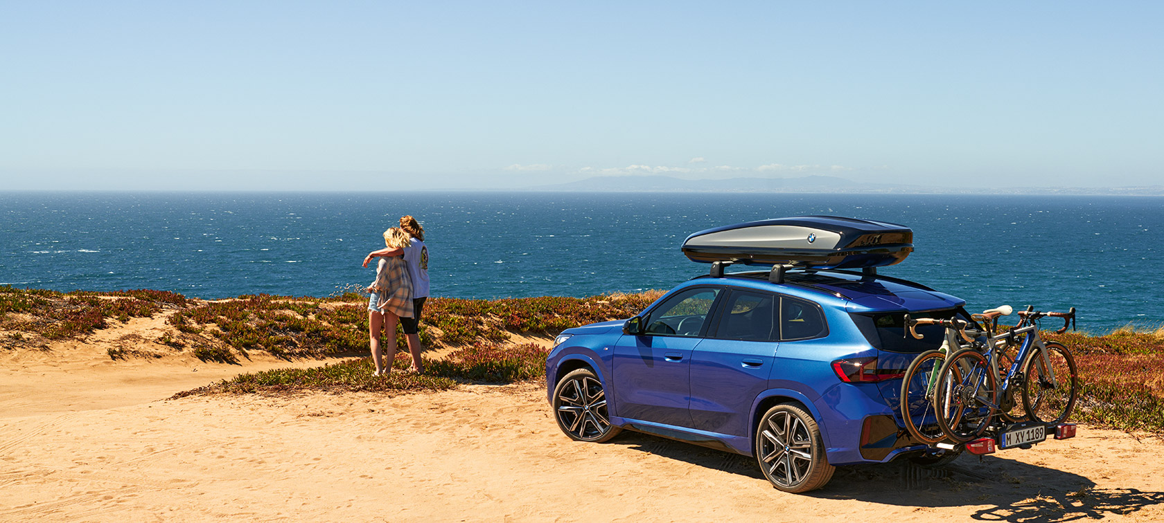BMW Originaltilbehør for vår og sommer BMW X1 U11 BMW Originaltilbehør for vår og sommer BMW X1 U11 på sand stående foran kysten med 2 personer