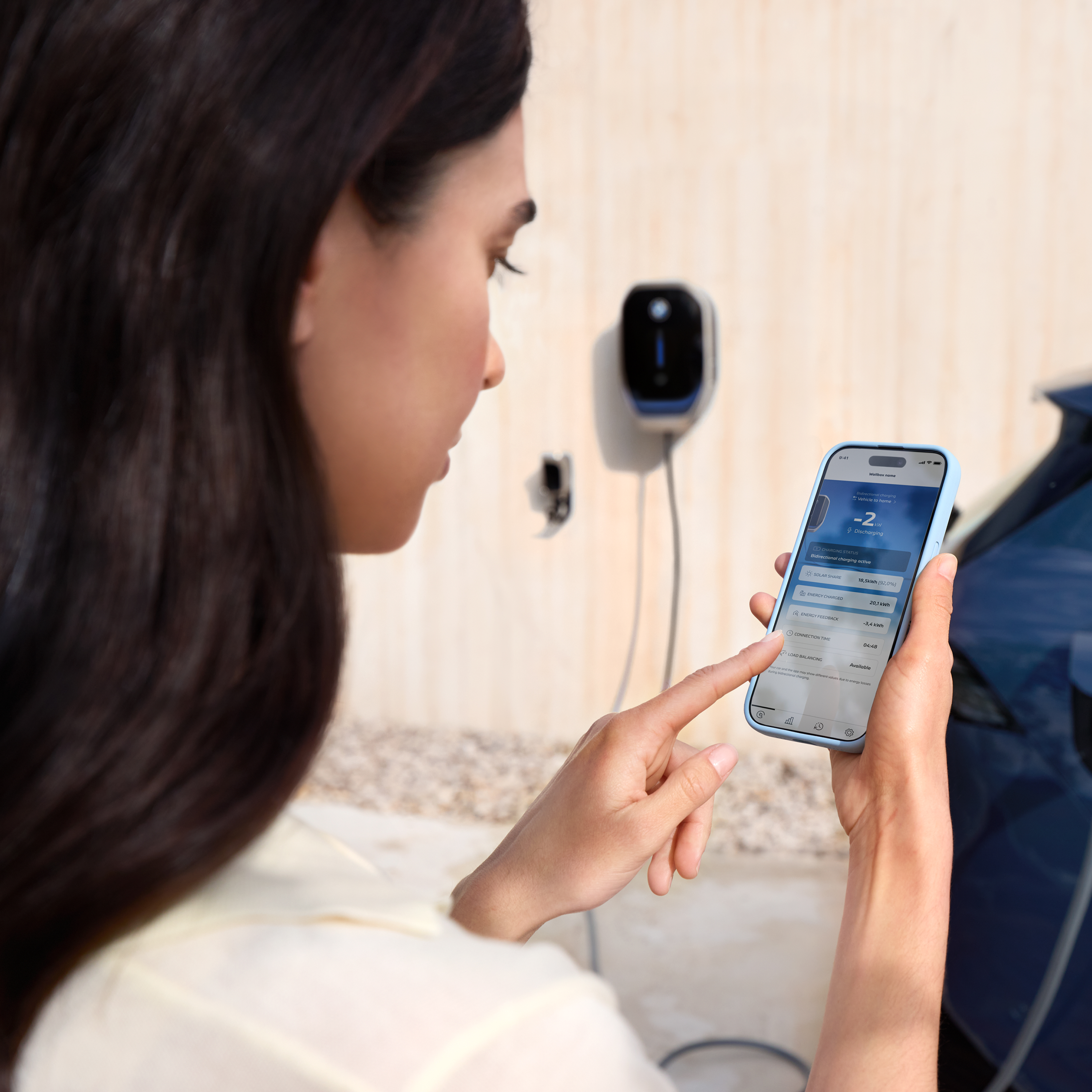 En kvinne ser på smarttelefonen sin, en BMW Wallbox er synlig i bakgrunnen